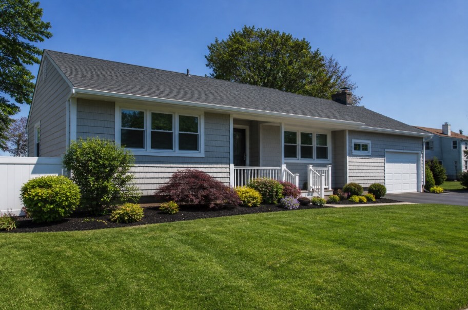 Wide view of a well-maintained single-story ranch home with gray siding, a manicured green lawn, and neatly landscaped mulch beds along the foundation under a clear blue sky.
